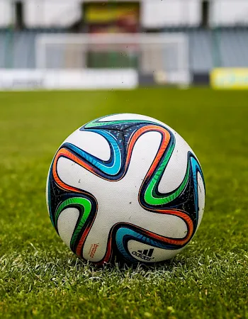 A close-up view of a colorful soccer ball resting on a green football pitch, with a blurred goal and stadium in the background.